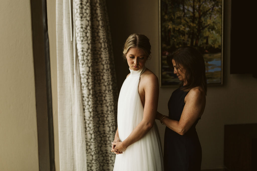 Bride stands in front of floor to ceiling window looking over left shoulder while mother fastens waist of wedding dress