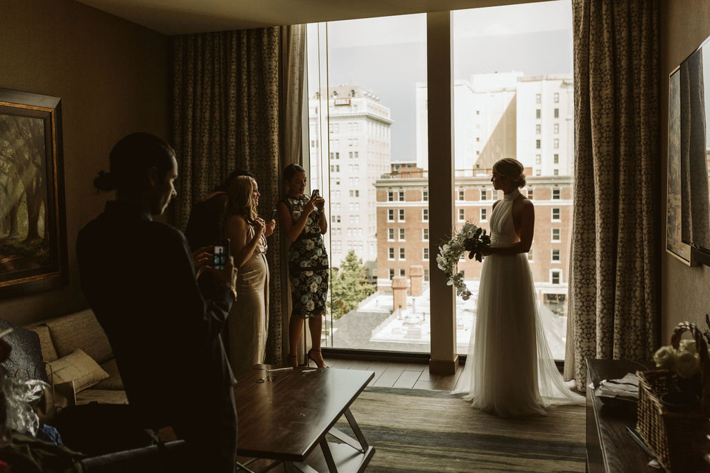 Bride with bouquet in front of floor-to-ceiling window overlooking downtown Chattanooga while friends take photos
