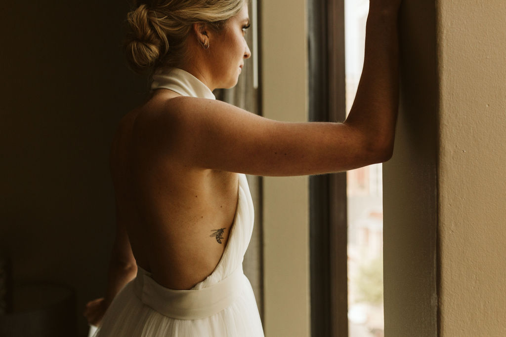 woman with bumblebee tattoo wearing backless wedding dress leaning into a window overlooking downtown Chattanooga