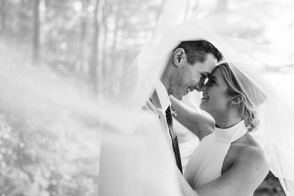 Couple cuddle nose-to-nose under a white veil, her hand on his shoulder