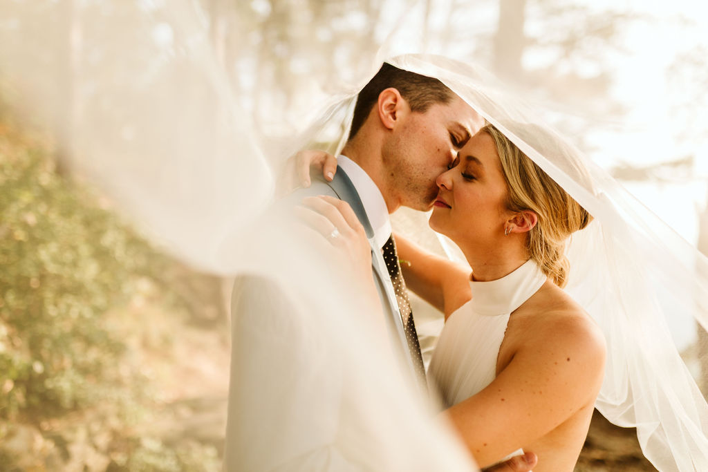 Groom kisses bride's cheek under a white veil, her hand on his shoulder