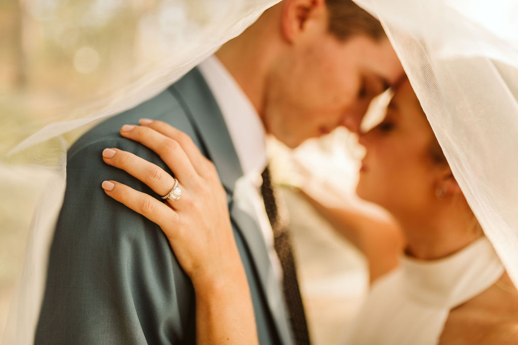 Close-up of bride's emerald cut diamond ring as bride and groom snuggle nose-to-nose under bride's veil