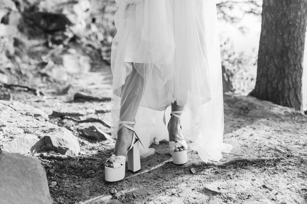 Bride stands with the hem of her dress pulled up to reveal her white stacked heels on a dirt trail