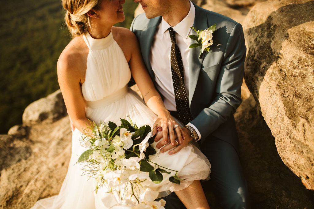 Bride in halter-style dress holding bouquet of white flowers sits with groom's hand on her knee