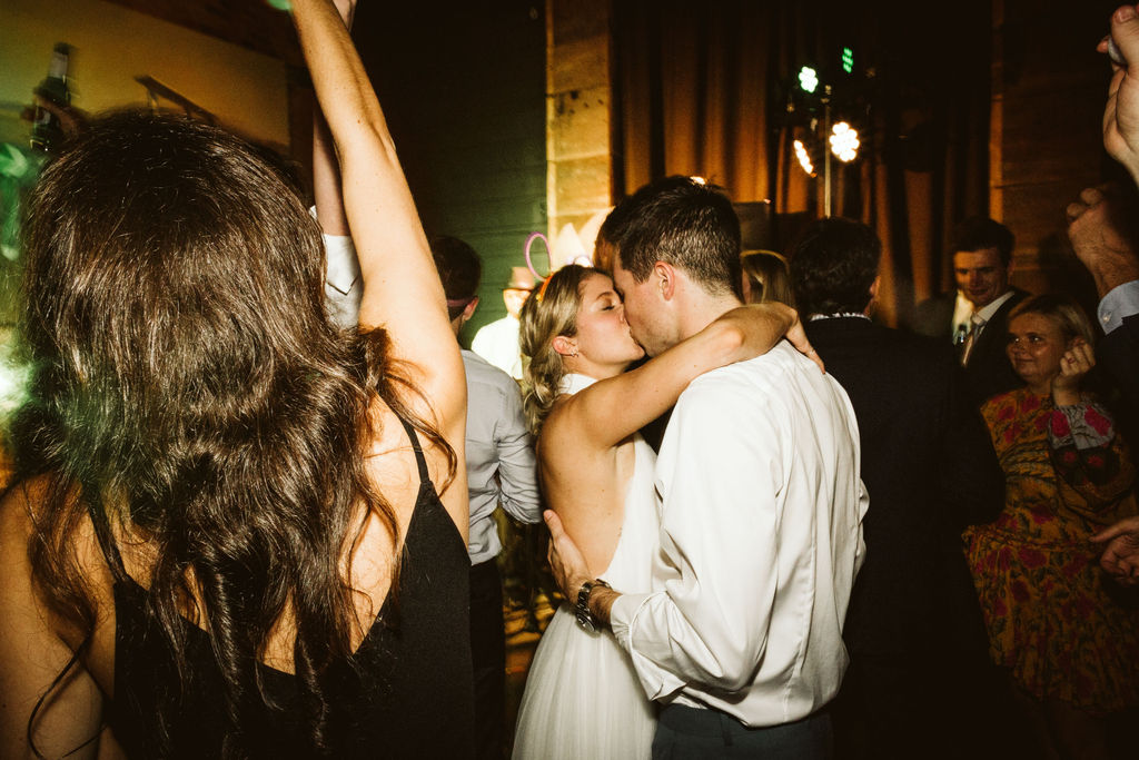 Bride and groom hold each other and kiss while guests dance around them