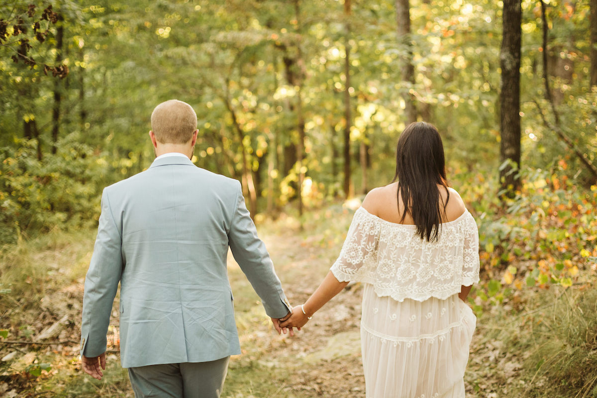 Man in light blue suit and woman in white lacy dress hold hands beneath tall trees