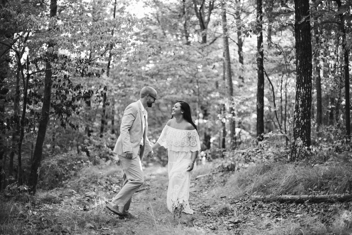 Man in light blue suit and woman in white lacy dress stand beneath tall trees