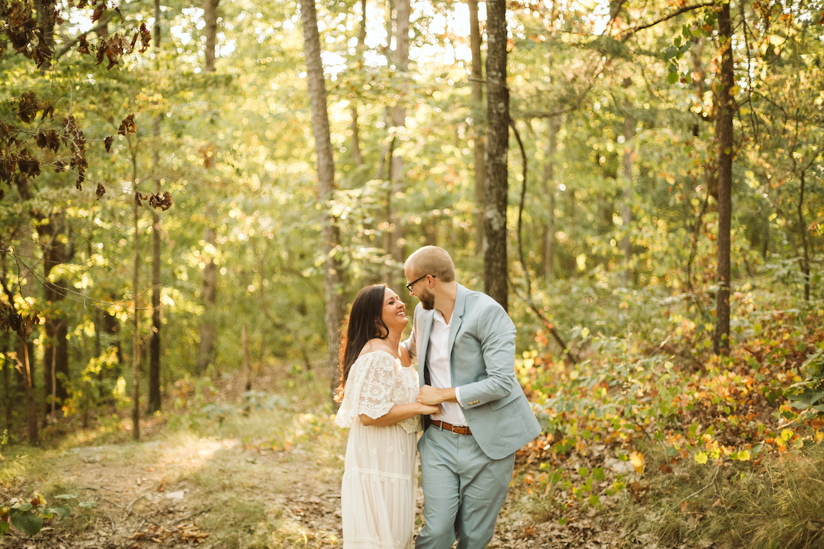 Man in light blue suit and woman in white lacy dress stand beneath tall trees