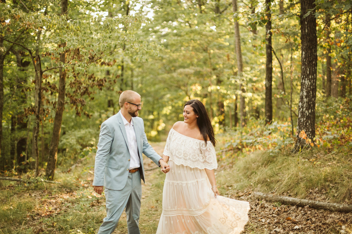 Man in light blue suit and woman in white lacy dress stand beneath tall trees