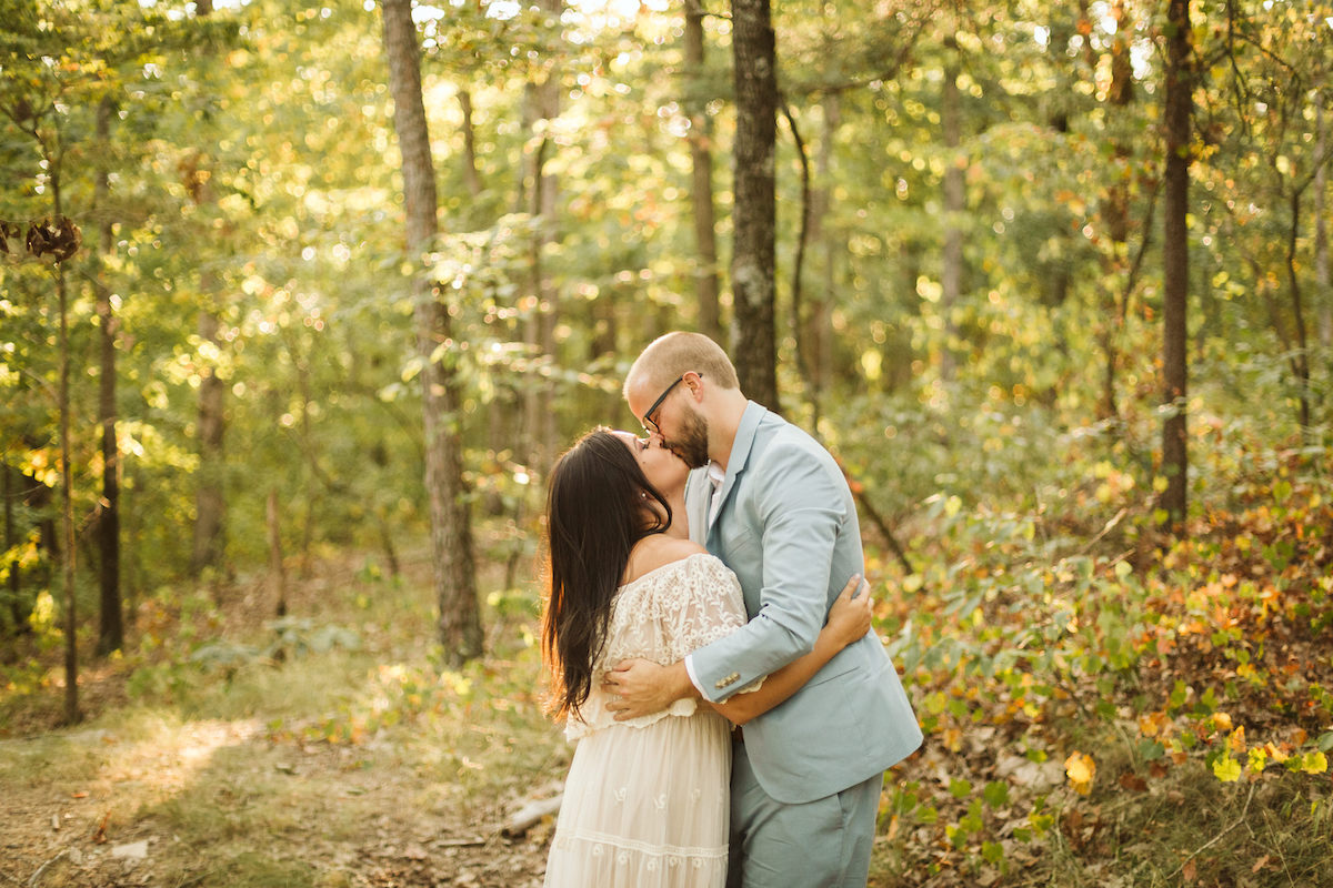 Man in light blue suit and woman in white lacy dress kiss beneath tall trees