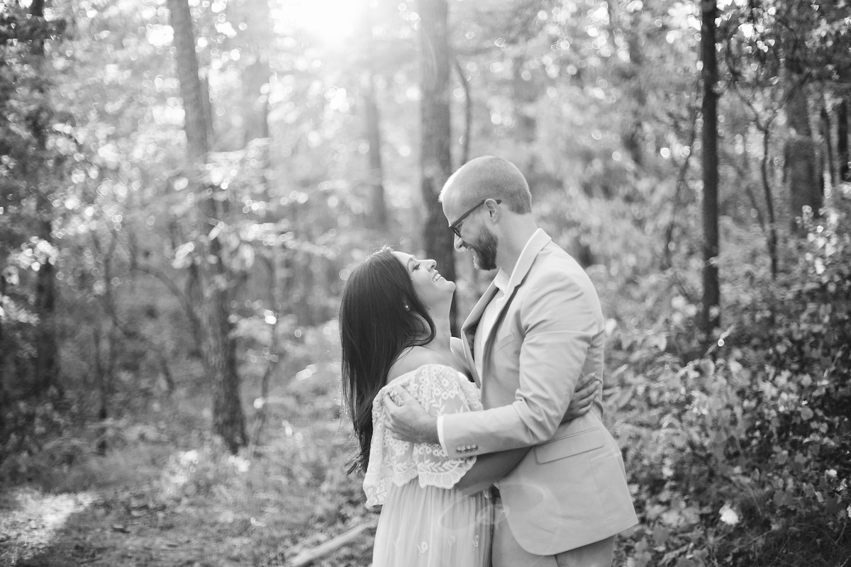 Man in light blue suit and woman in white lacy dress stand hugging, looking at each other beneath tall trees