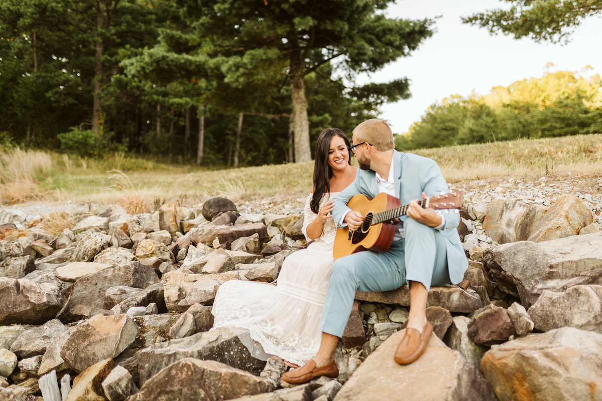 Man in light blue suit and woman in strapless white dress sit on large rocks near woods. He plays a guitar while she smiles.