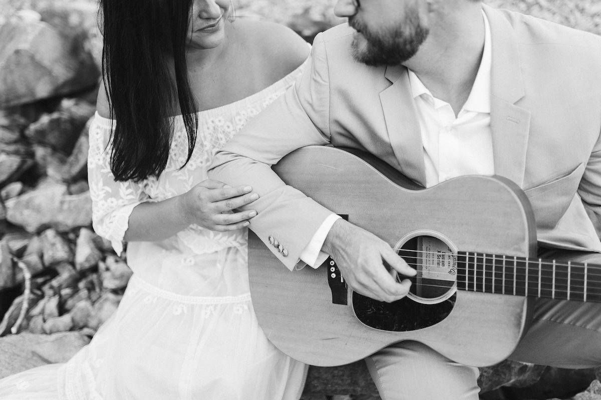 Man in light blue suit and woman in strapless white dress sit on large rocks. He plays a guitar while she smiles.