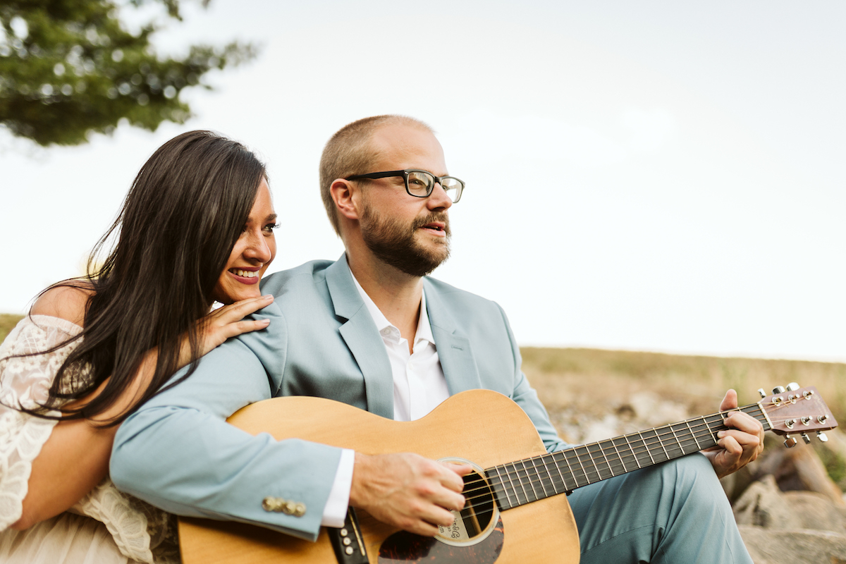 Man in light blue suit and woman in strapless white dress sit on large rocks. He plays a guitar while she smiles.