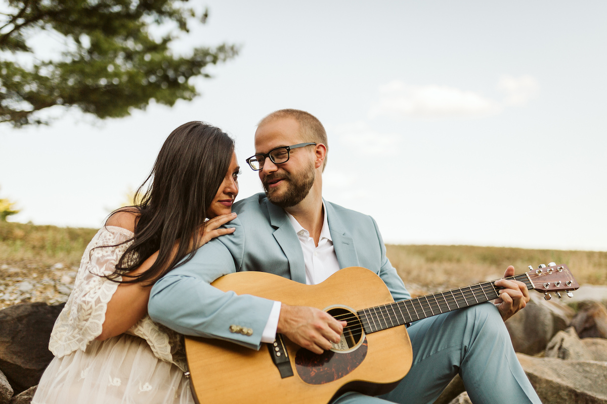Man in light blue suit and woman in strapless white dress sit on large rocks. He plays a guitar while she smiles.