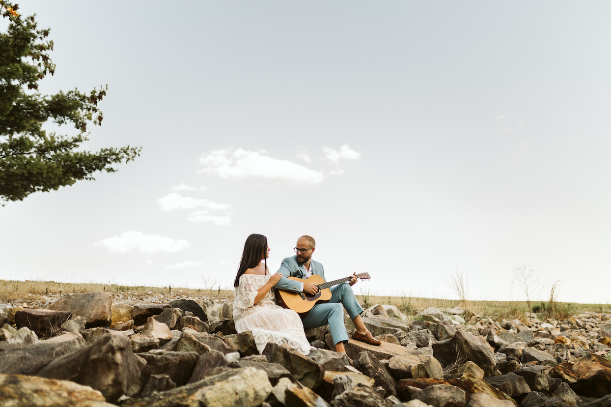 Man in light blue suit and woman in strapless white dress sit on large rocks. He plays a guitar while she smiles.