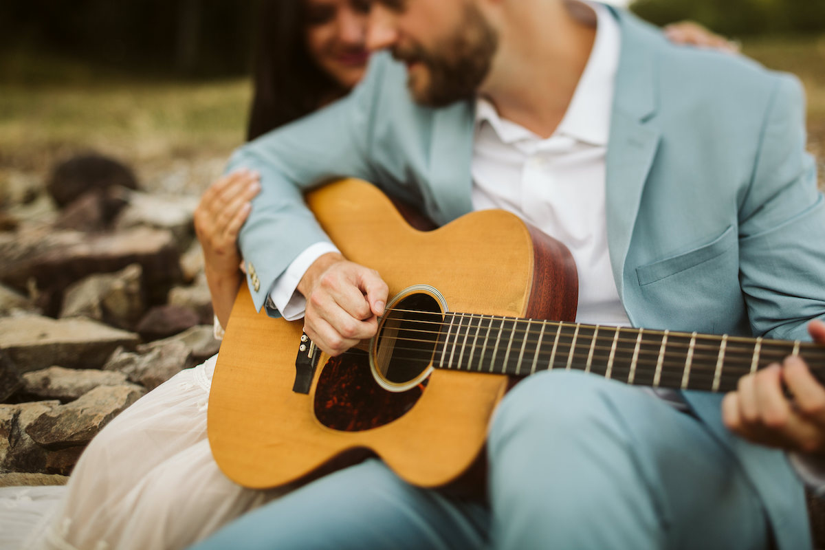 Man in light blue suit and woman in strapless white dress sit on large rocks. He plays a guitar while she smiles.