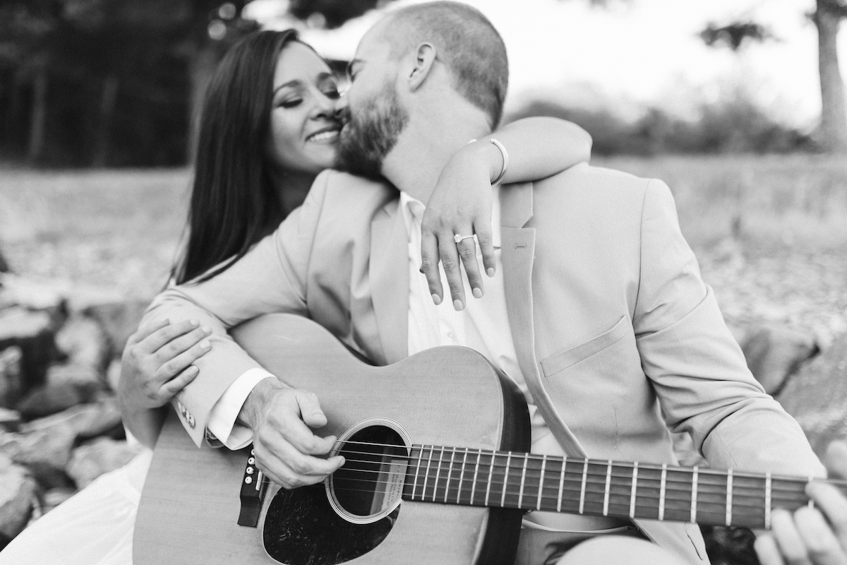 Man in light blue suit and woman in strapless white dress sit on large rocks. He plays a guitar while she smiles.