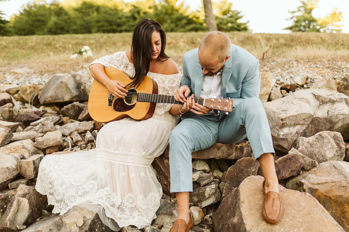 Man in light blue suit and woman in strapless white dress sit on large rocks. She plays a guitar as he reaches over the frets