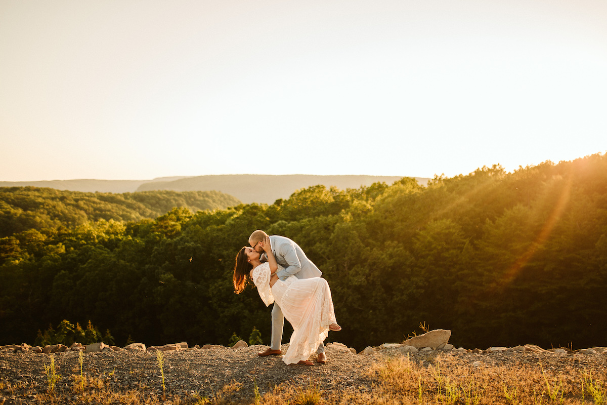 Man in light blue suit dips woman in flowing lacy white dress at sunset with rolling Tennessee Valley hills behind them