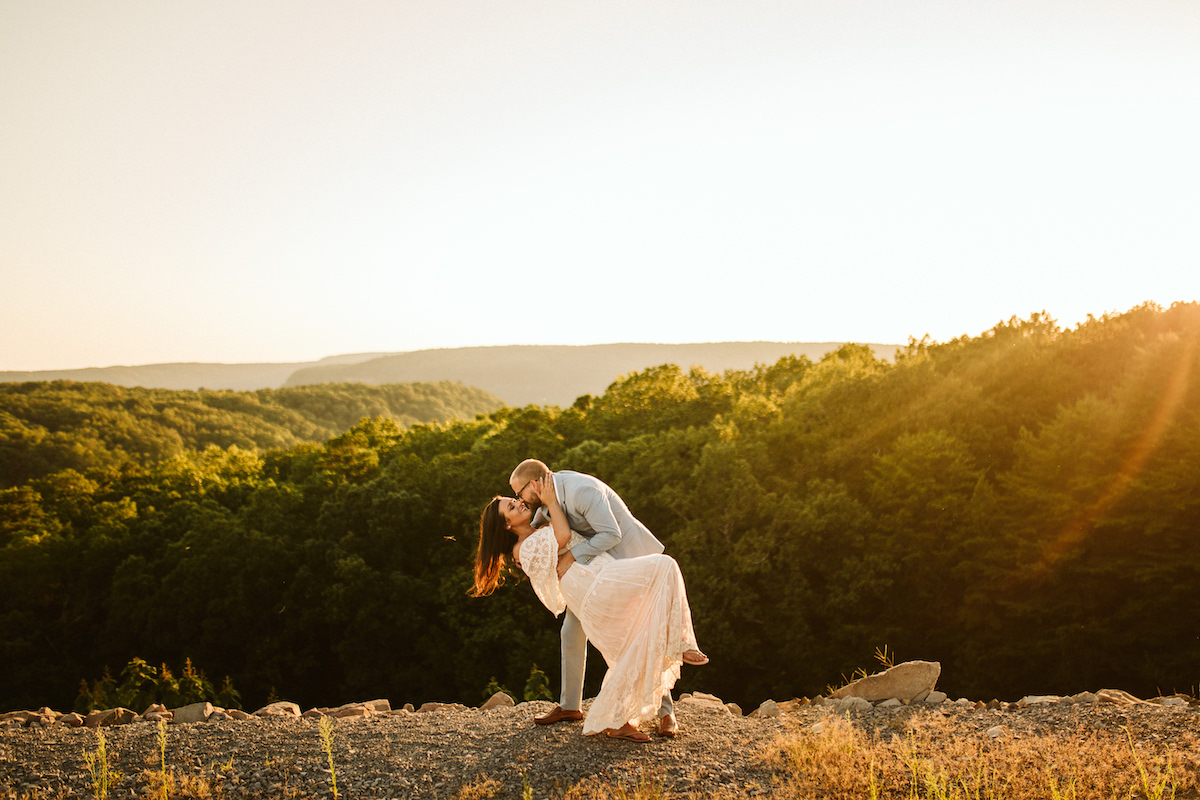 Man in light blue suit dips woman in flowing lacy white dress at sunset with rolling Tennessee Valley hills behind them