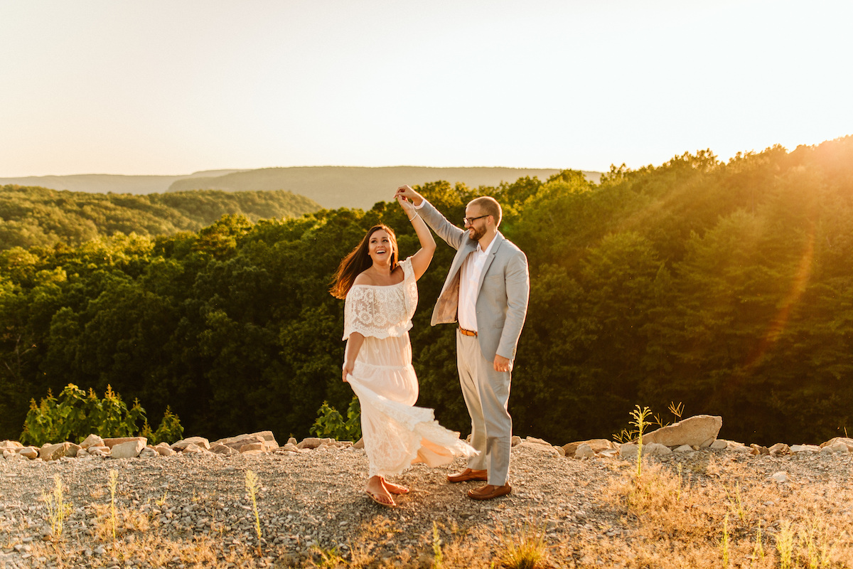 Man in light blue suit twirls woman in flowing lacy white dress at sunset with rolling Tennessee Valley hills behind them