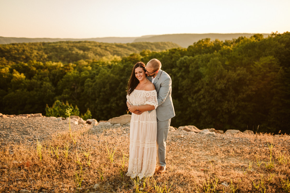 Man leans over woman's shoulder, almost kissing her neck while they both smile. Behind them the sun sets over several hills.