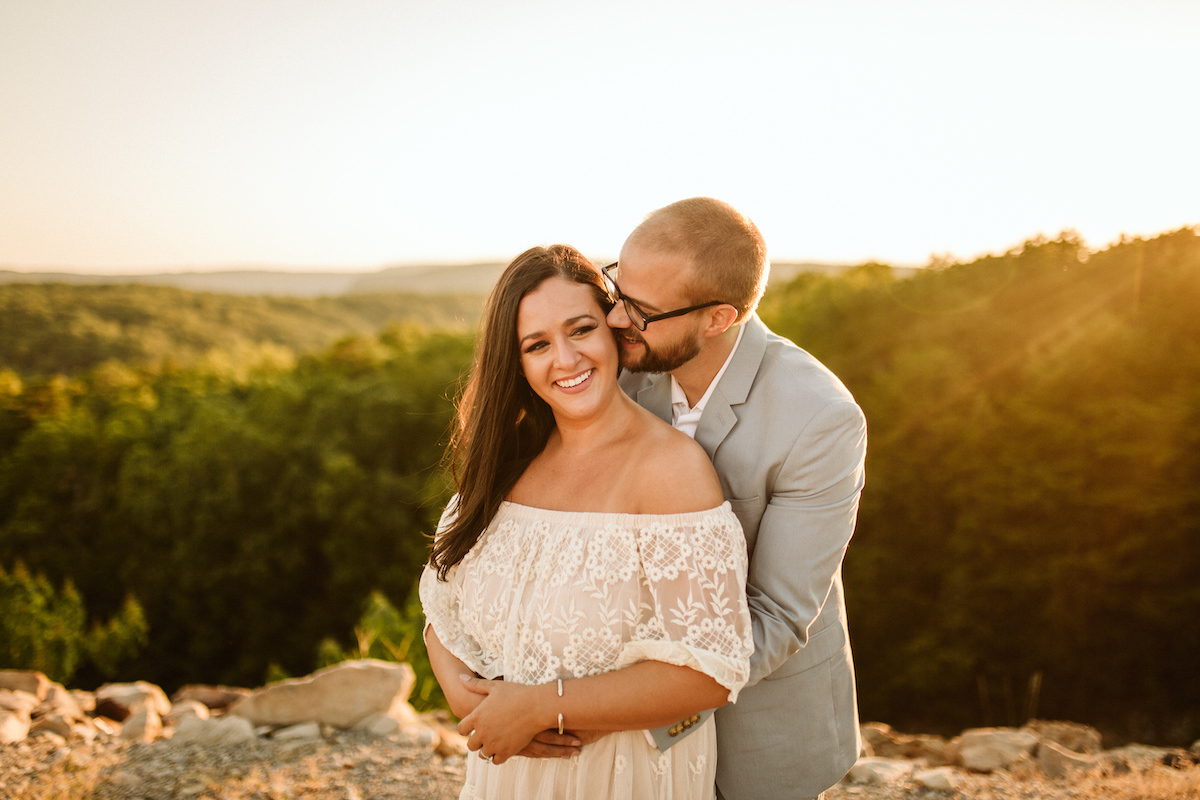 Man leans over woman's shoulder, almost kissing her neck while they both smile. Behind them the sun sets over several hills.