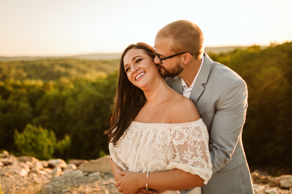 Man leans over woman's shoulder, almost kissing her neck while they both smile. Behind them the sun sets over several hills.