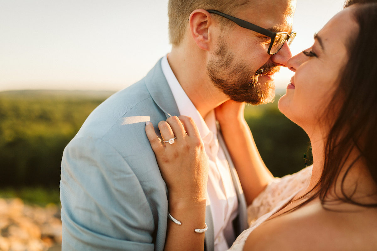 Man in light blue suit and glasses leans nose-to-nose with woman at sunset during Raccoon Mountain engagement session