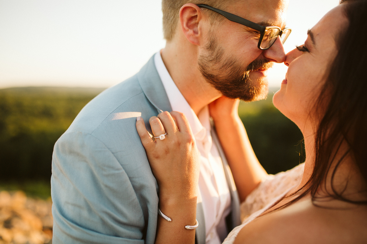 Man in light blue suit and glasses leans nose-to-nose with woman at sunset during Raccoon Mountain engagement session
