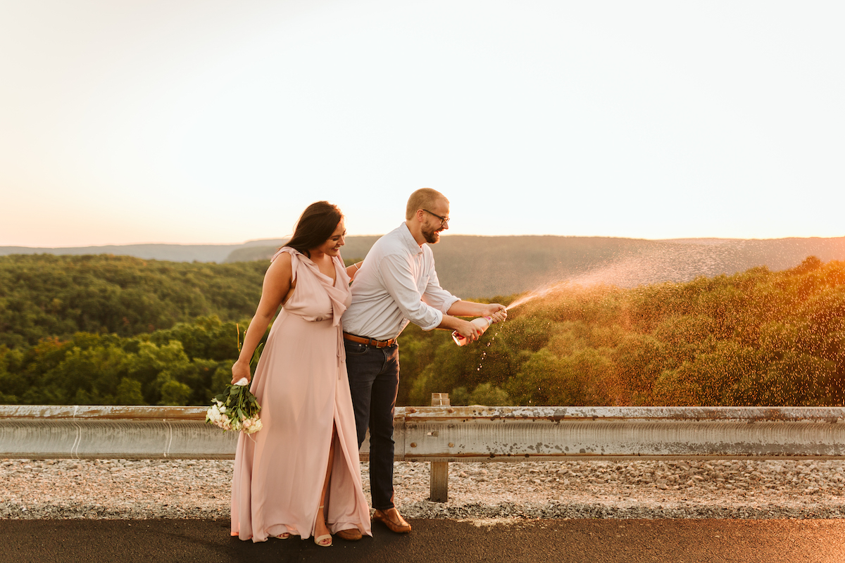 Man uncorks champagne and champagne sprays out. Woman in pink dress stands next to him laughing.