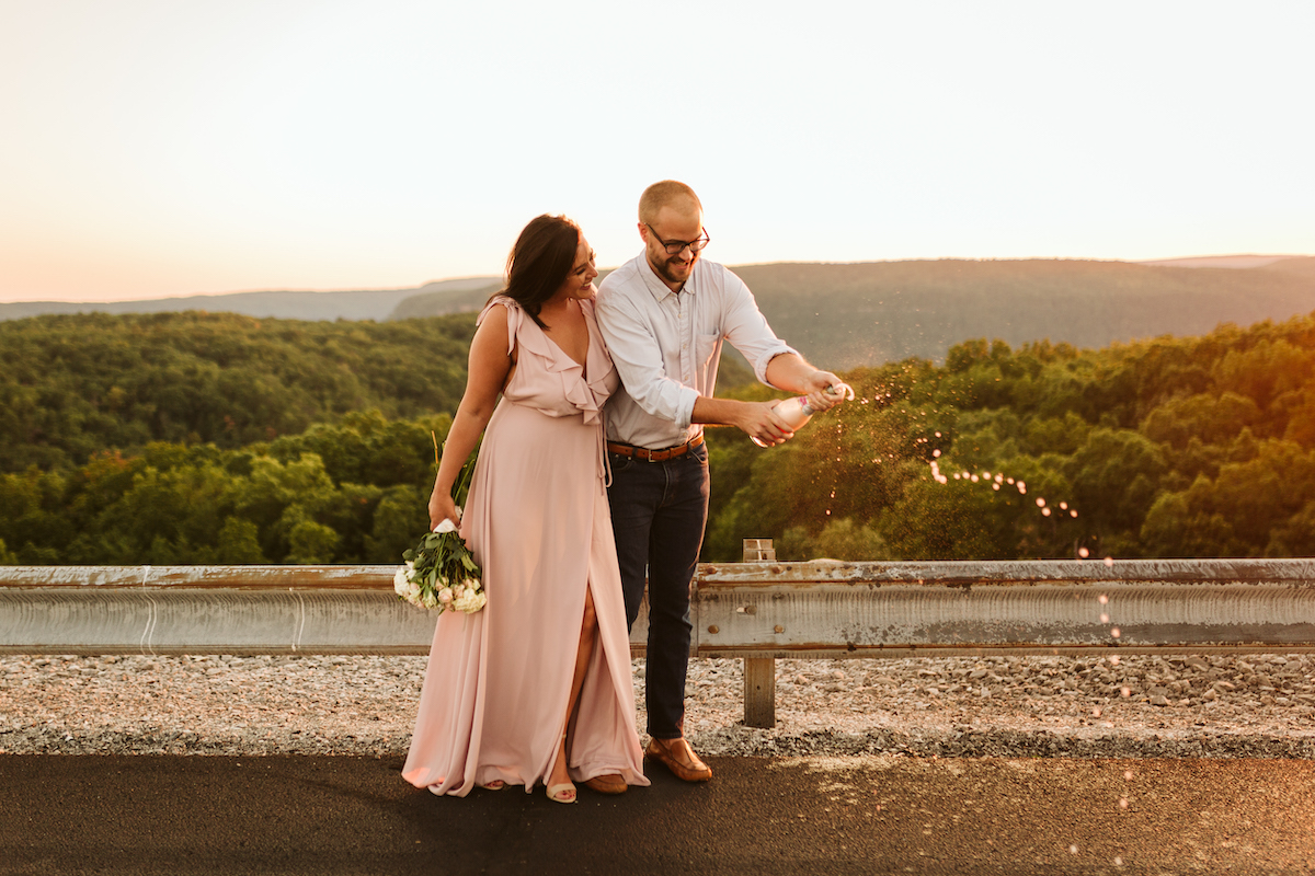 Man uncorks champagne and champagne sprays out. Woman in pink dress stands next to him laughing.