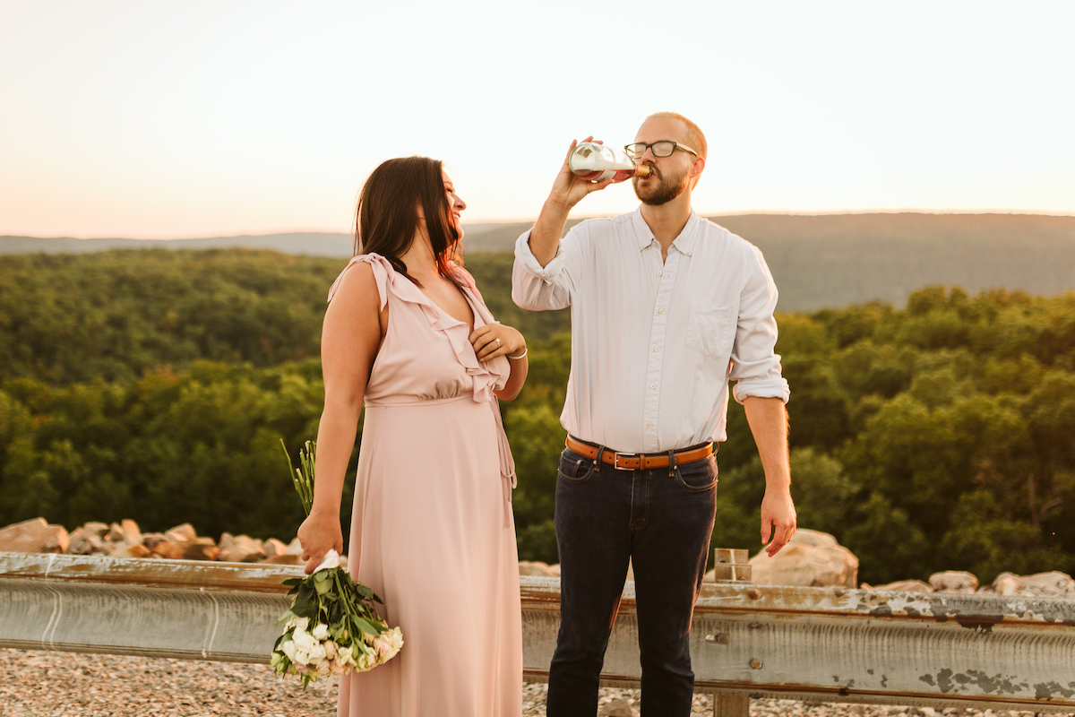 Man drinks champagne from the bottle. Woman in pink dress stands next to him laughing.