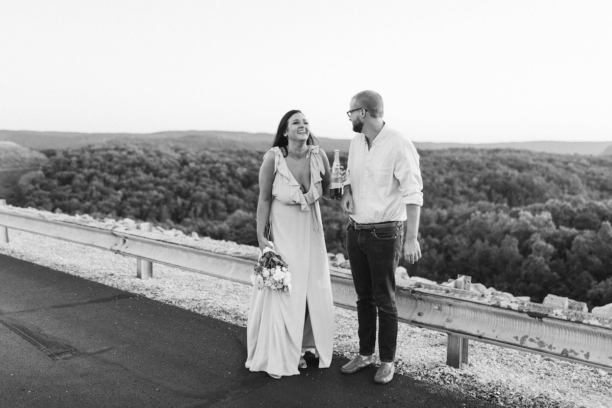 Man and woman stand next to each other on paved road near metal guardrail. She holds champagne bottle and laughs.