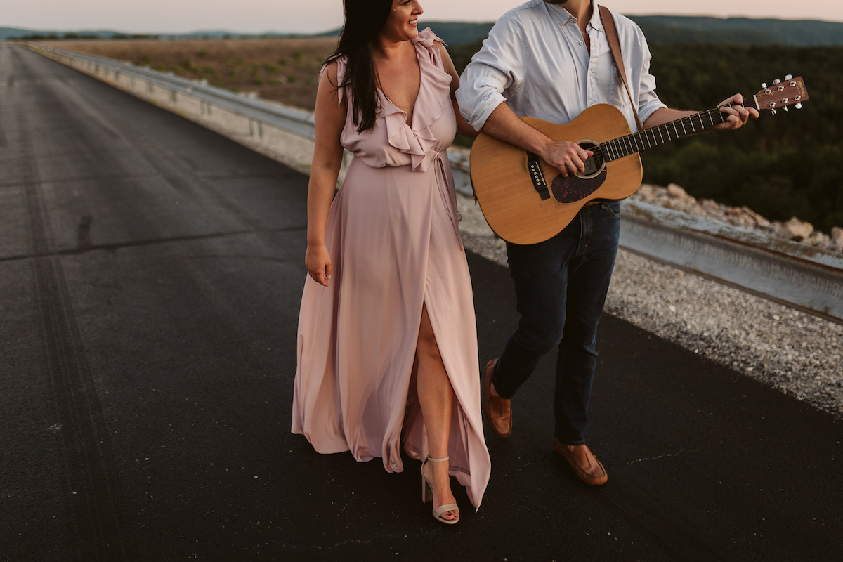 Man and woman walk on paved road at golden hour. He strums a guitar hung over his shoulder.
