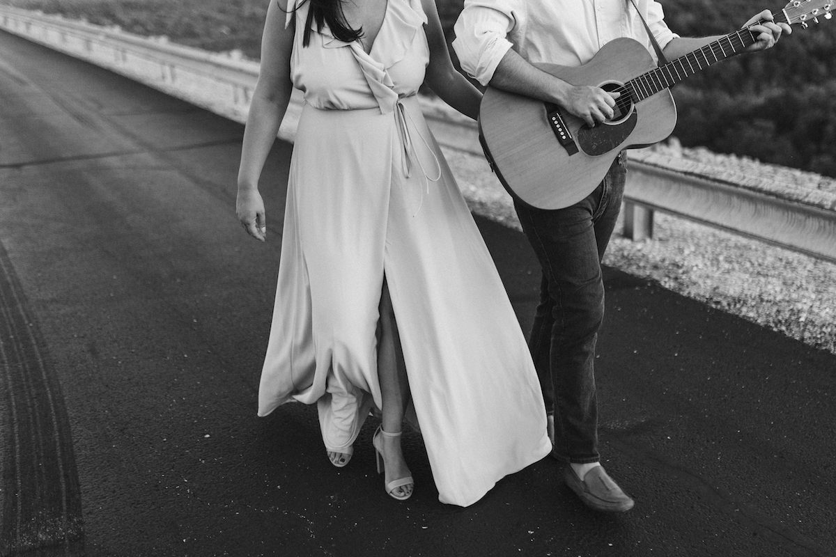 Man and woman walk on paved road. He strums a guitar hung over his shoulder.