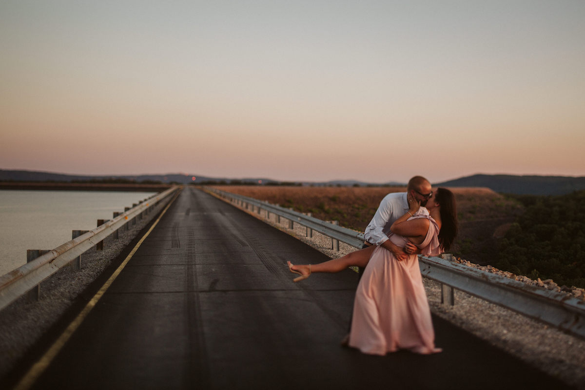 Man dips woman in long pink dress back and kisses her at golden hour on Raccoon Mountain's loop drive