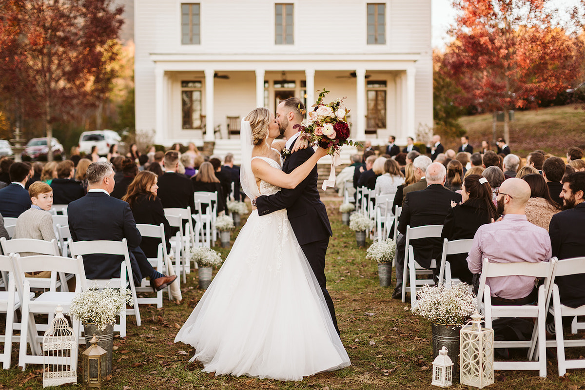 Bride and groom hug and kiss at the end of the aisle with The Homestead at Cloudland Station's Peacock Hall behind them