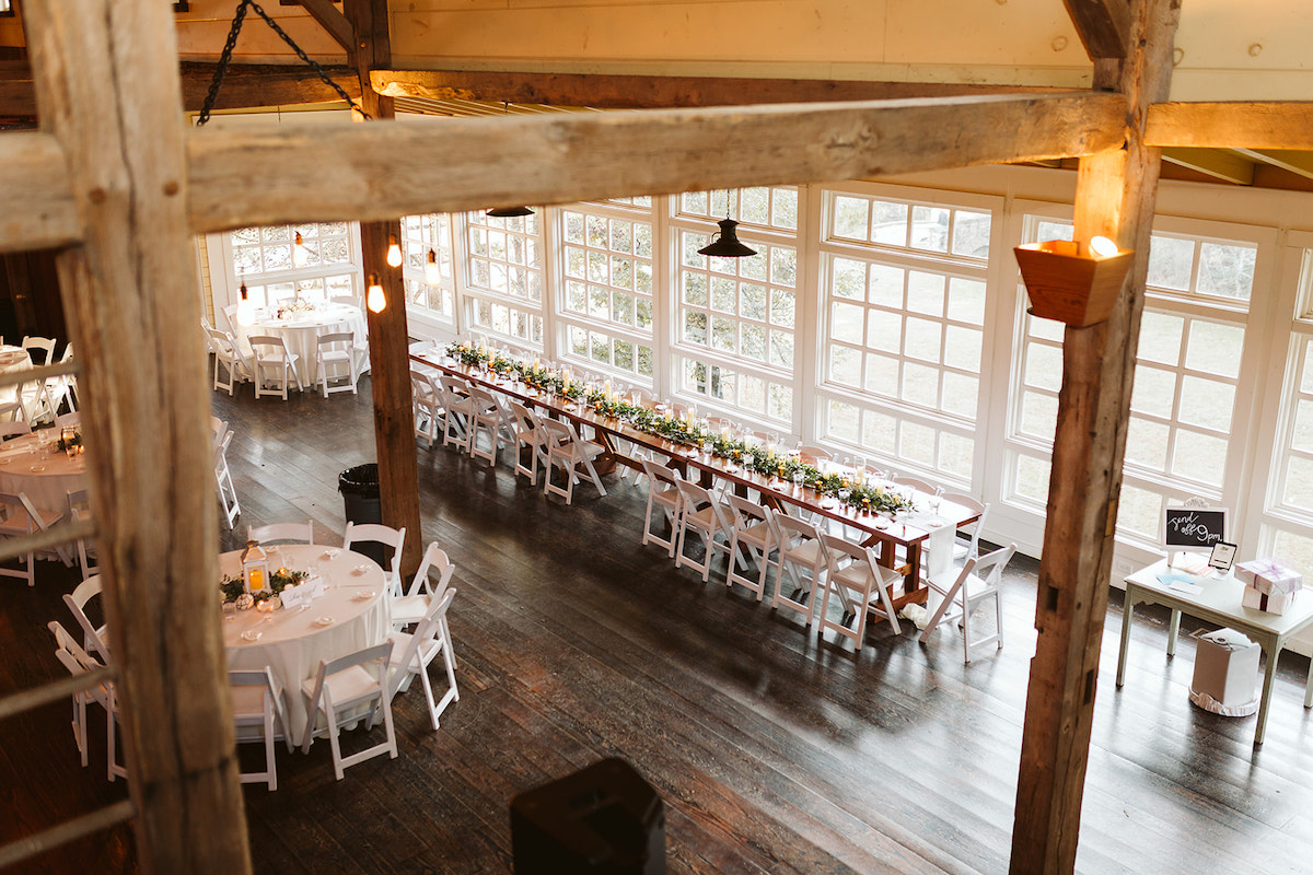 Long, farmhouse table in front of a wall of white windows under the high, open rafters of a barn's interior