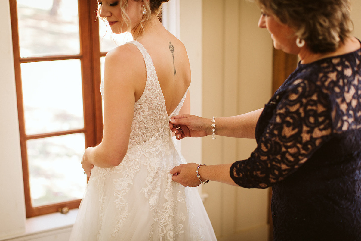 Mother in dark, lacy dress, zips the back of bride's low-back lacy wedding dress