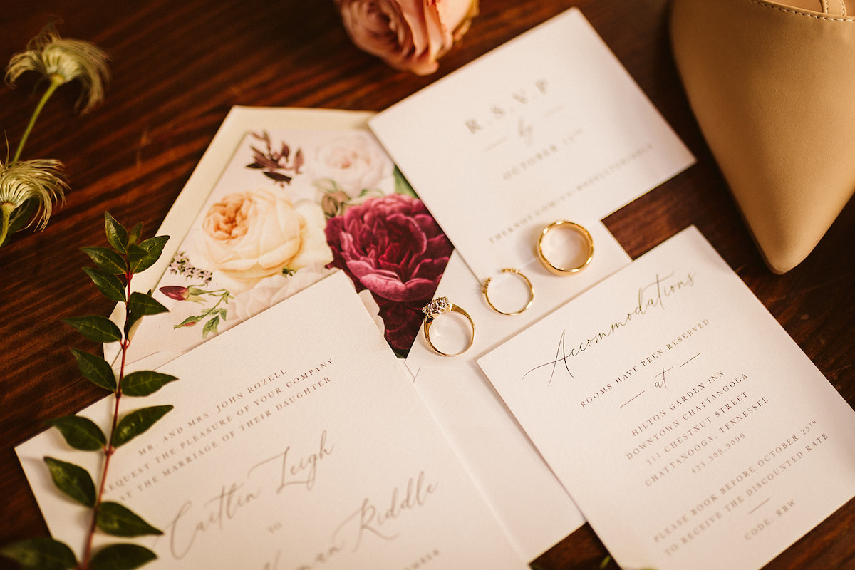 Wedding invitation and RSVP card lie between flowers and bride's shoes. The wedding bands sit on top of the cards.