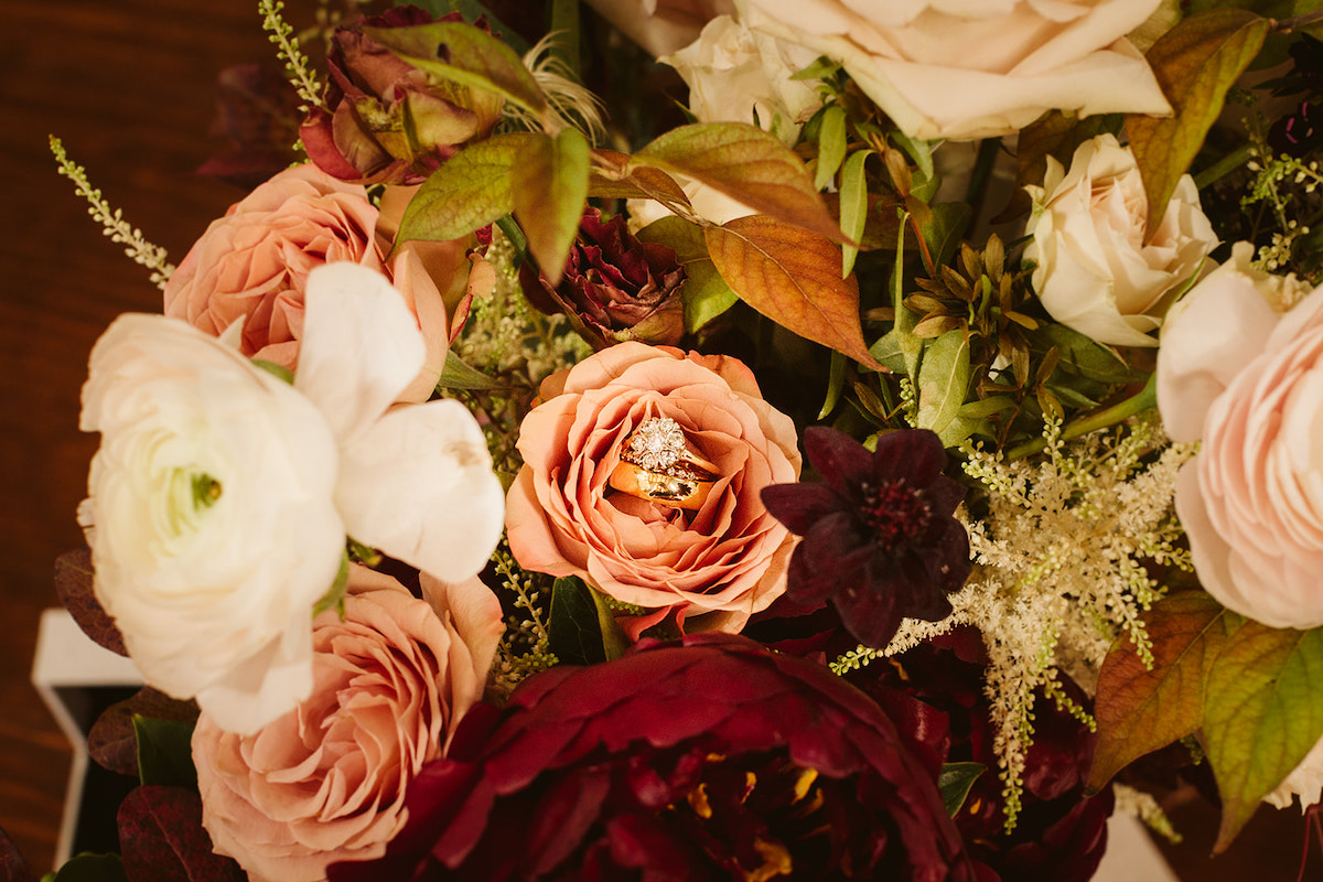 Wedding bands nestled inside the petals of a peach rose. Dark red flowers and greenery make up the rest of the bouquet.