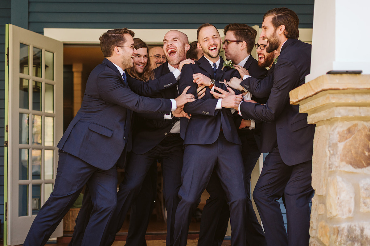 Groomsmen gather around the groom on a porch, pushing him and laughing. They all wear dark suits and brown leather shoes.