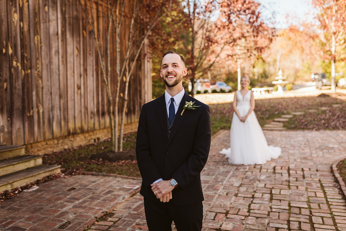 Groom stands on a wide brick carriage path next to a tall barn. His bride stands behind him on the path before the First Look