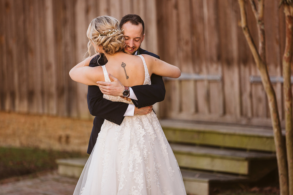 Groom hugs bride in front of dark barn. Her low-back lacy dress reveals a black key tattoo on her upper back.