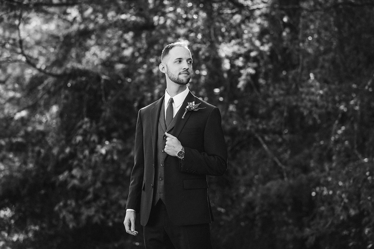 Groom wears a dark suit over a gray vest, navy tie and white shirt. He holds his lapel and his watch peeks out of his sleeve.