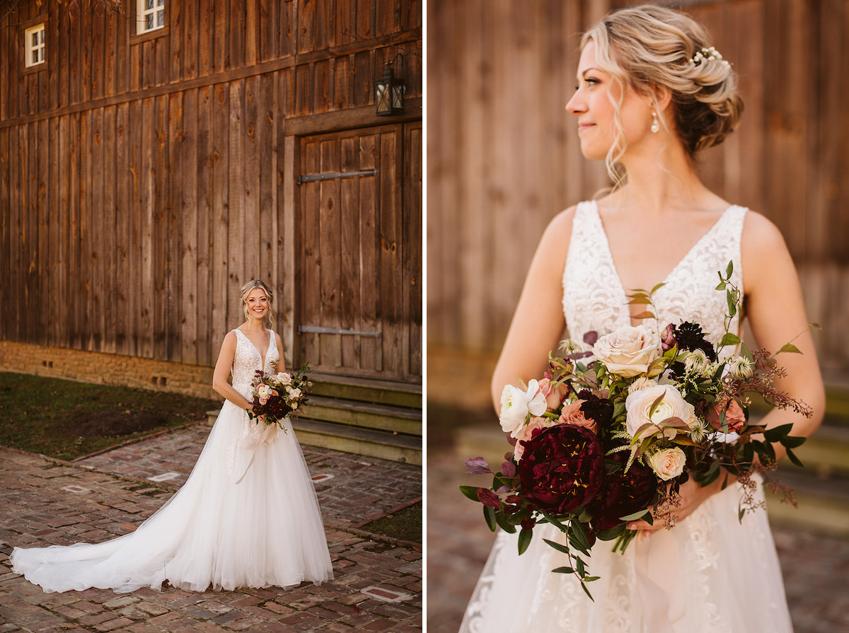 Bride holds bouquet of deep red, peach, and white flowers with greenery. She stands on a brick path next to a tall wooden barn.
