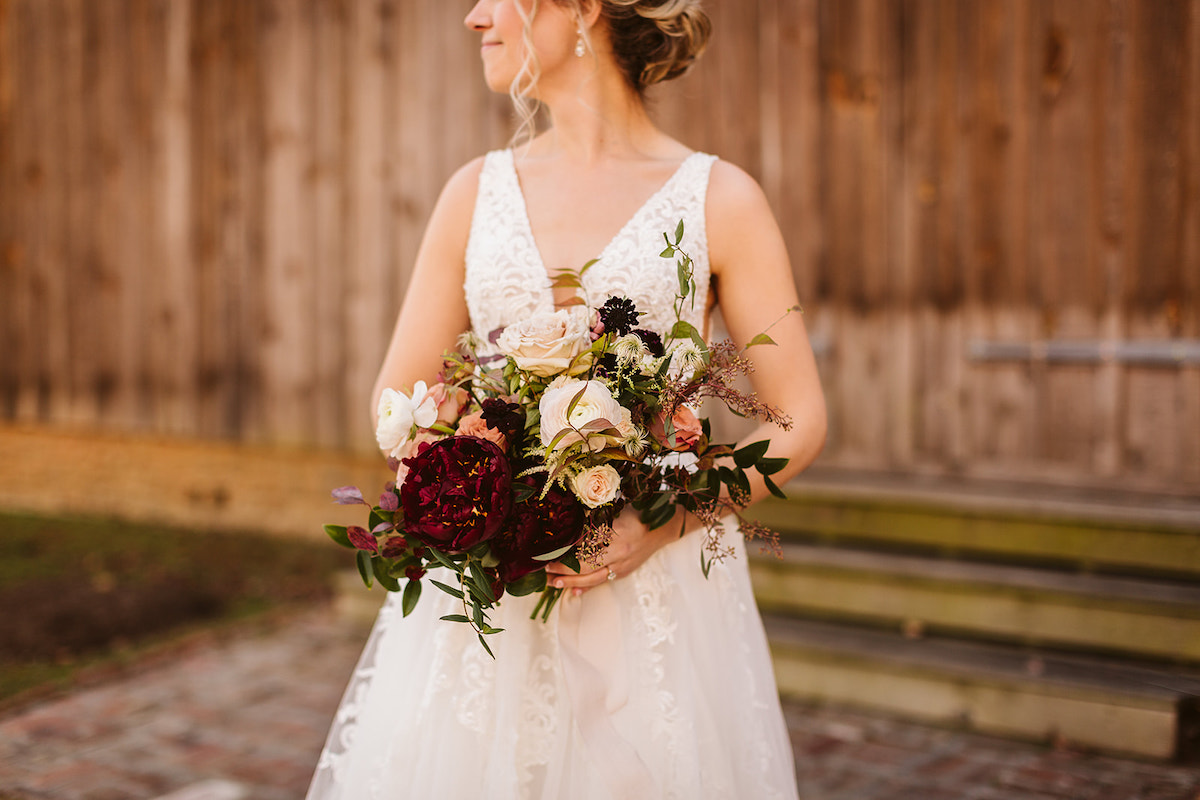 Bridal holds her Petaline bouquet filled with dark red flowers, pink and white roses, and greenery against her lace gown.