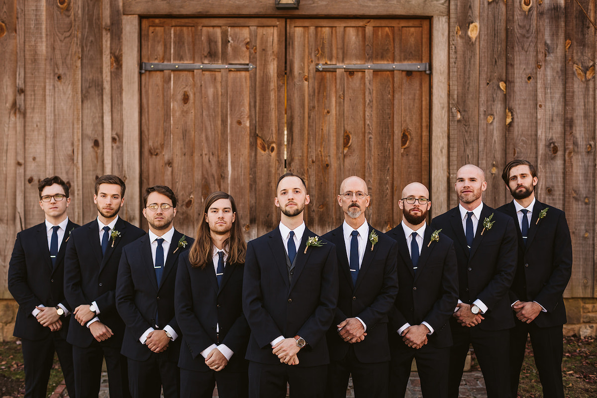 Groom stands with groomsmen in their dark suits in front of tall wooden barn doors